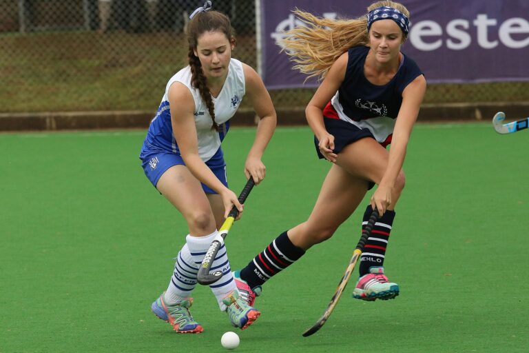 Two female field hockey players in action on a vibrant green pitch.